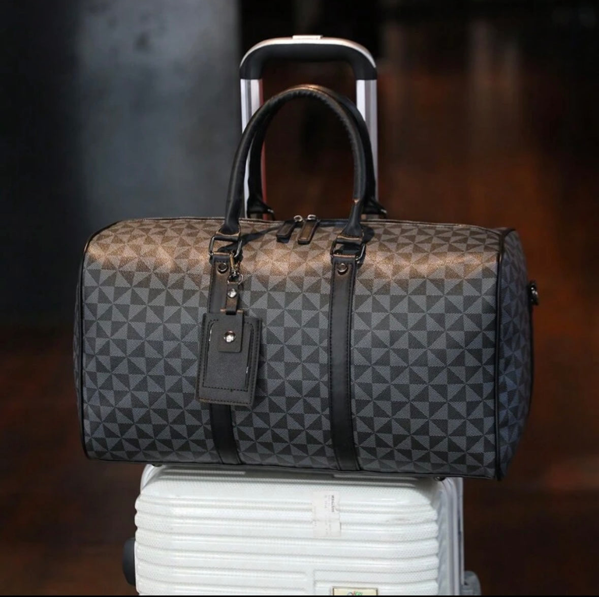 A stylish black and gray patterned duffle bag resting on top of a white rolling suitcase. The bag has two leather handles, black straps, and a luggage tag attached, making it suitable for travel.